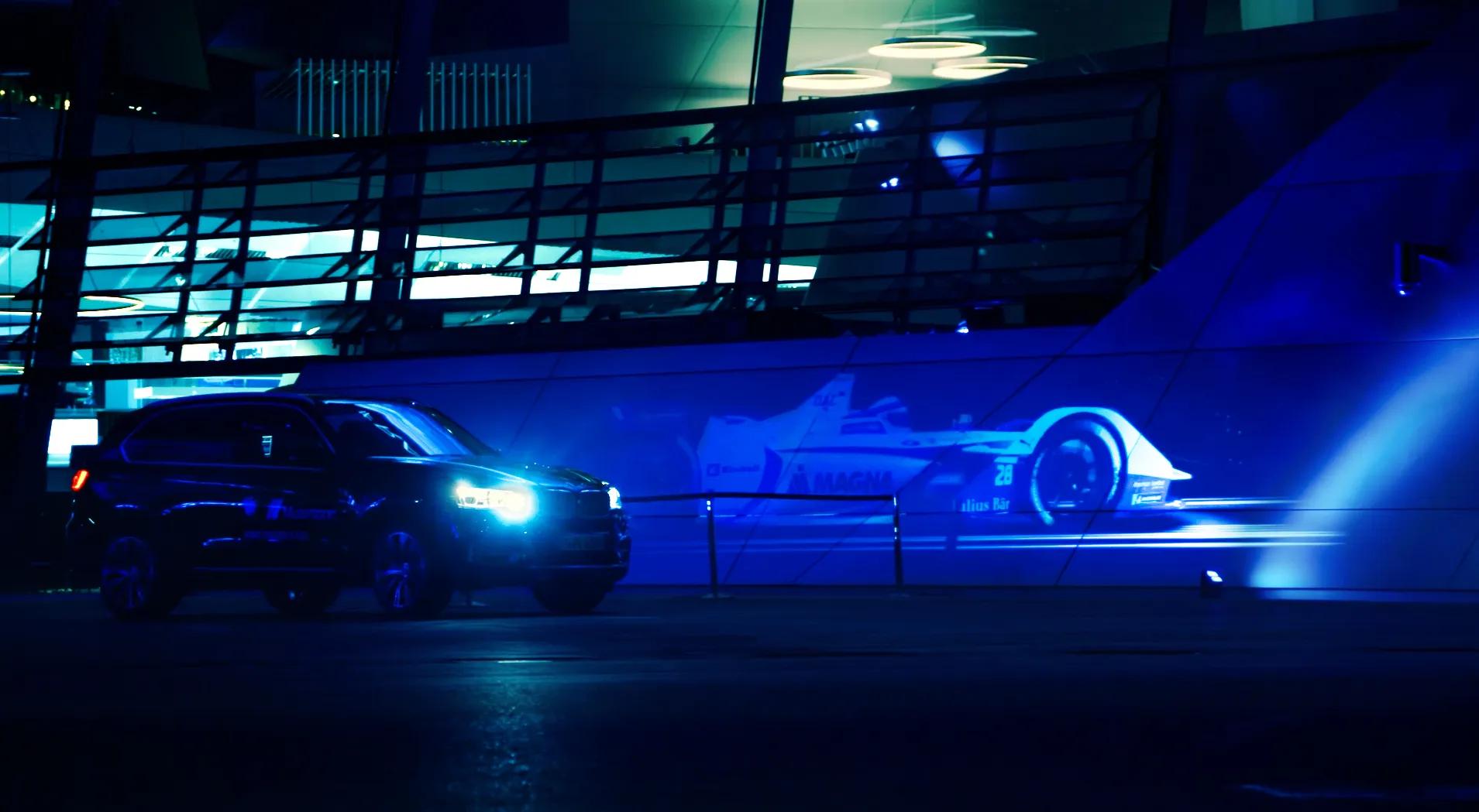 Formula E race car projected onto a building wall beside a BMW at night