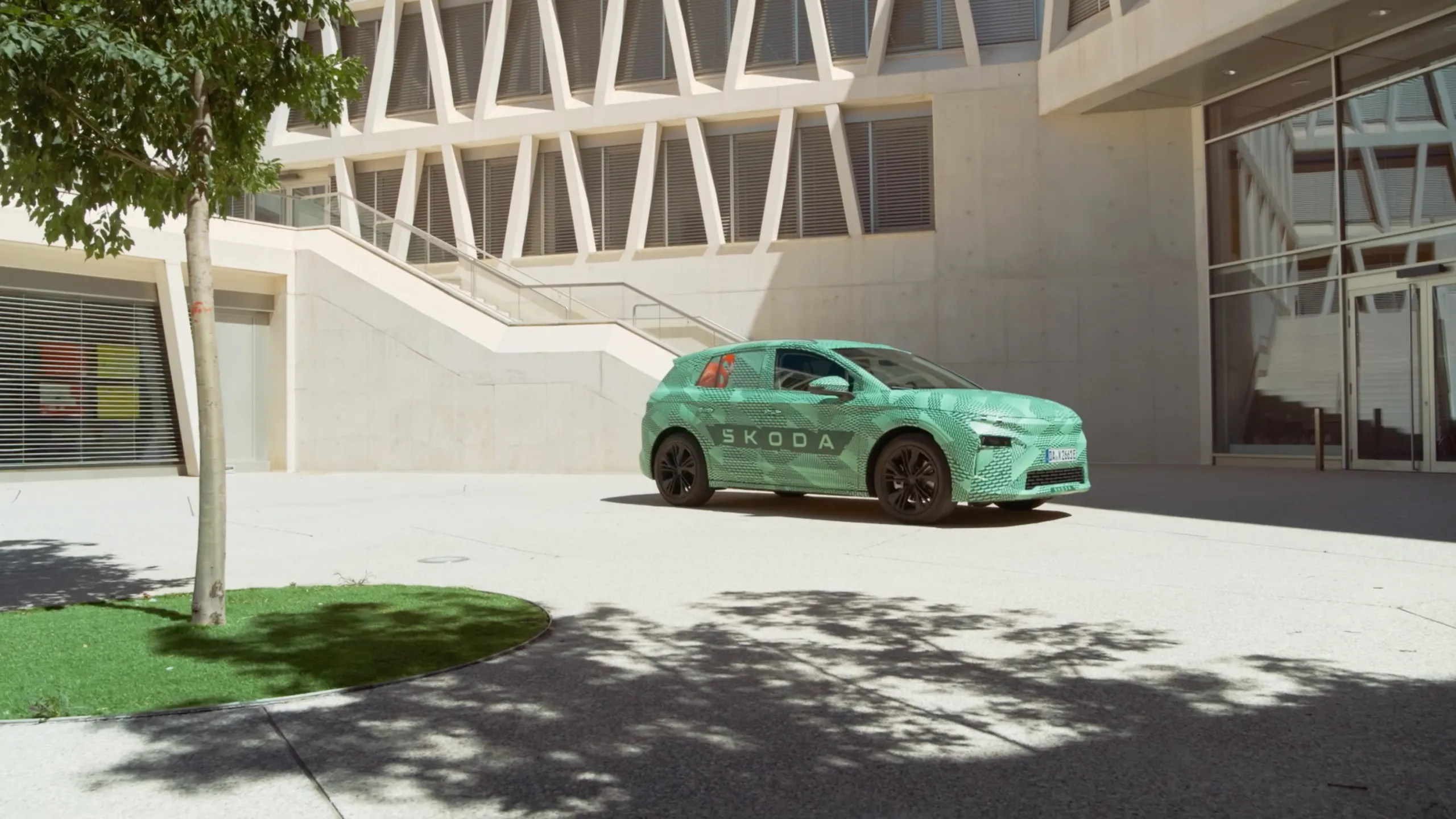 Camouflaged Škoda Elroq parked in sunlight in front of modern concrete architecture
